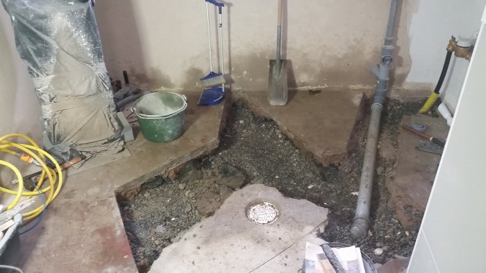 Uppgrävt källargolv med avloppsrör och makadam runt ett större hål.