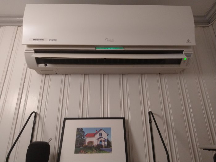 Rengjord innedel av en luftvärmepump monterad på vägg ovanför en inramad bild av ett hus.