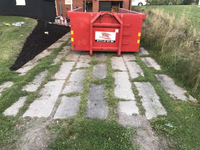 Röd container för blandat byggavfall framför hus, med rensad yta med bark till vänster och ogräs till höger.