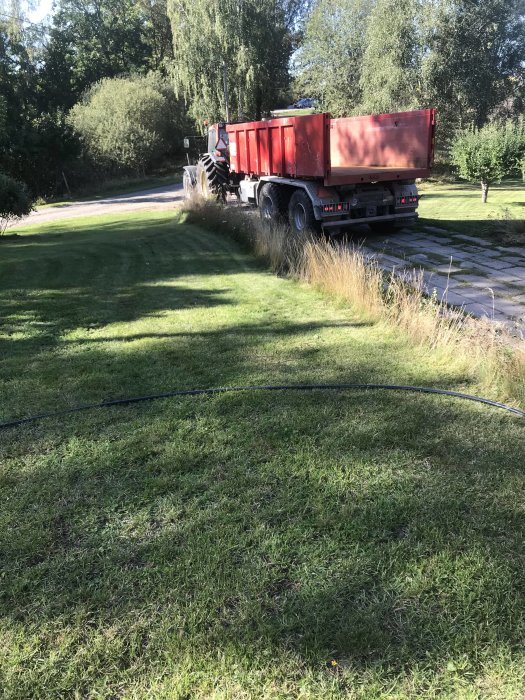 Röd container på släpkärra parkerad på gräs vid uppfart med högt ogräs på ena sidan.