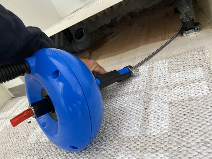 Person som använder en blå kabelinföringshjälp för att dra en kabel under golvet via en öppning vid en vägg.