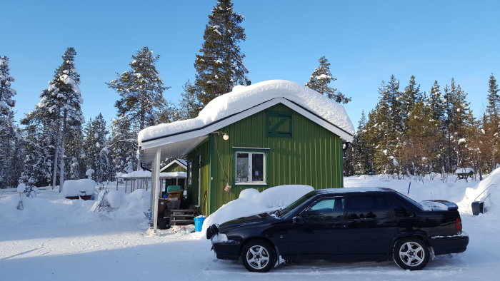 Ett grönt hus och en svart bil täckt med snö omgivna av snöklädd skog under en klar vinterdag.