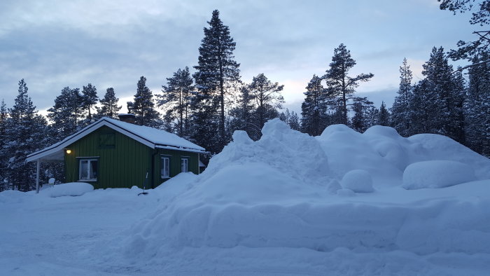 Grönt hus med snötäckt tak och stora snödrivor framför i en skogstomt vid vintertid.