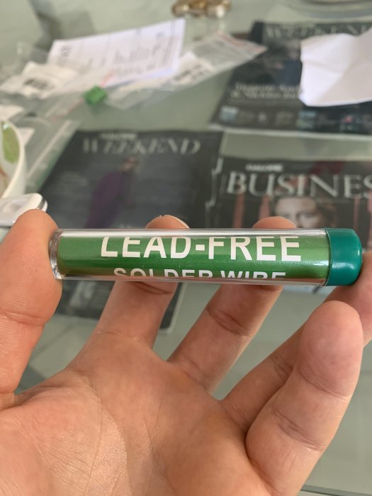 Hand håller lödtråd med text "LEAD-FREE SOLDER WIRE" framför rörig bakgrund.