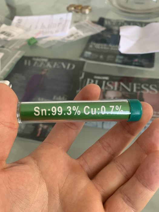 Hand håller en lödrulle med texten "Sn:99.3% Cu:0.7%", vilket indikerar en blyfri lödlegering.