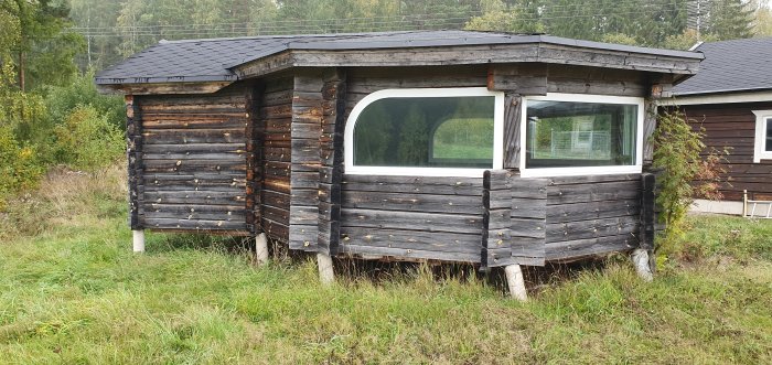 Ett litet timrat uthus med svarta shingeltak och stort fönster mot en skogsbakgrund och gräs framför.