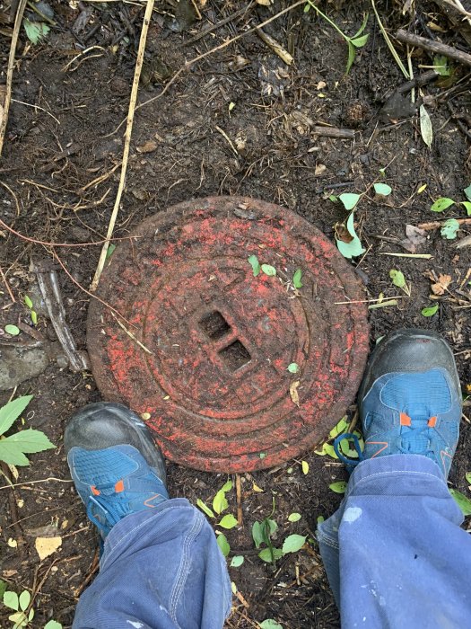 Skor som står framför en jordig, rostig brunnslock omgiven av vegetation.