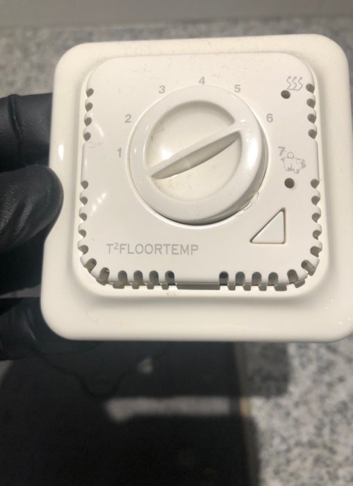Hand i svart handske håller en gammal givare med reglerknopp märkt "T2FLOORTEMP" för temperaturstyrning av golvvärme.