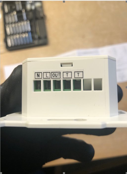 Termostatdosans insida med märkningar N, L, OUT, T och tomma anslutningar, hålls upp framför en verktygslåda.