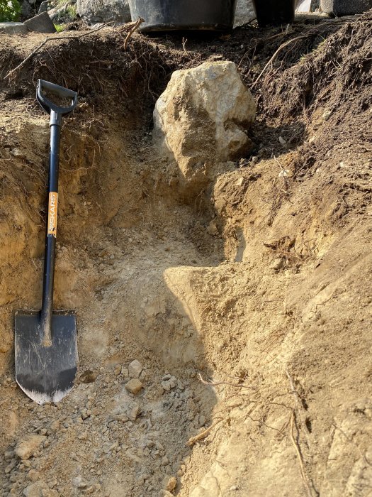 Grävd grop med en spade, stora stenar och synlig gjuten betongplatta i ett trädgårdsprojekt.