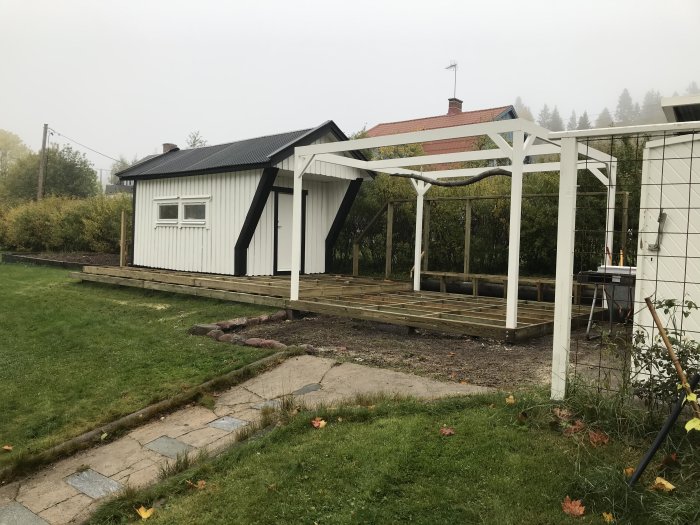 Nybyggt trädäck med pergola vid vit husfasad, fönsterbyte synligt, omgiven av grönska i disigt väder.