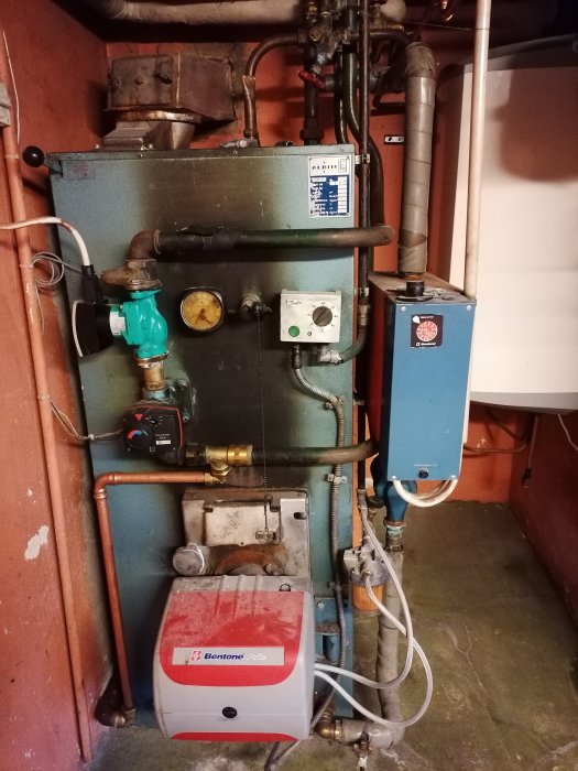 En äldre vattenburen uppvärmningspanna i ett pannrum märkt "Bentone", omgiven av rörkopplingar och ventiler.