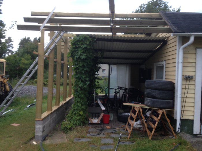 Nybyggd carport med reglar och takstomme kopplad till garage, omgiven av grönska.