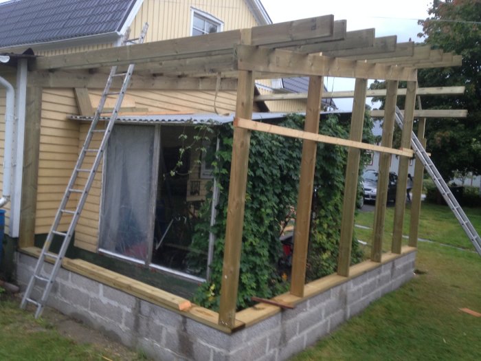 Halvfärdig träkonstruktion av en carport/grillplats vid ett hus, med lecablock och stegar.