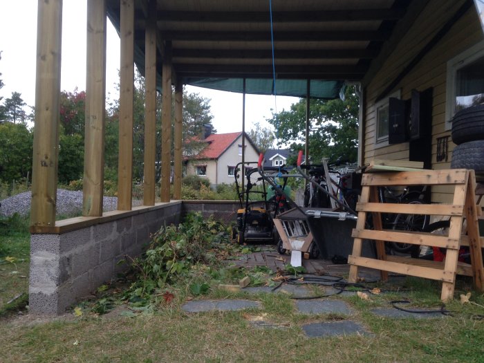 Nybyggd carport i anslutning till garage med murad grund och trästolpar, halvfärdig och med byggmaterial synligt.