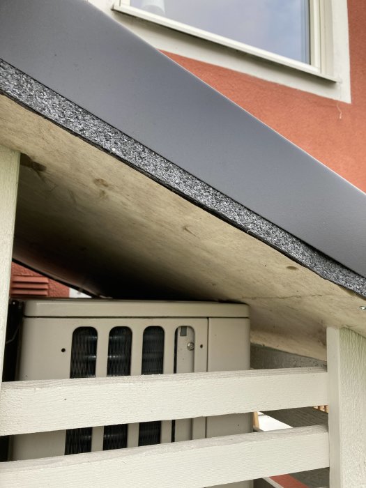 Ett skydd med tak över en utomhusdel till en luftvärmepump vid en husvägg, sett från snedvinkel ovanifrån.