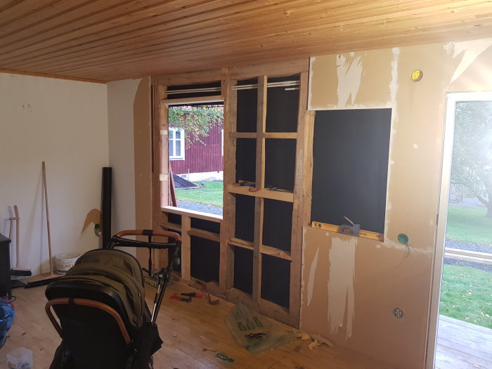 Inre vägg av en stuga under renovering med ett borttaget fönster och verktyg synliga.