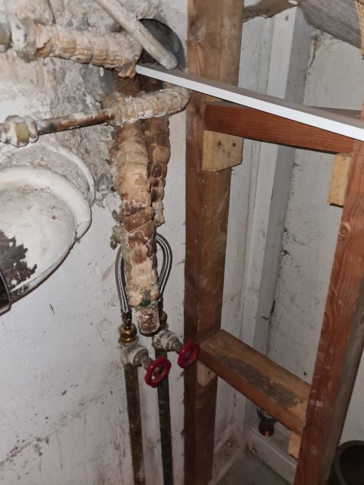 Rostig gammal stam med avlägsnad glasullisolering, kvarvarande rör med röda ventiler i en källare under renovering.