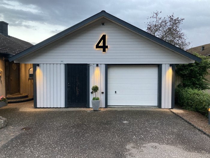 Renoverad vit garagedörr på en villa med nummer 4 och upplysta vägglampor i skymningen.