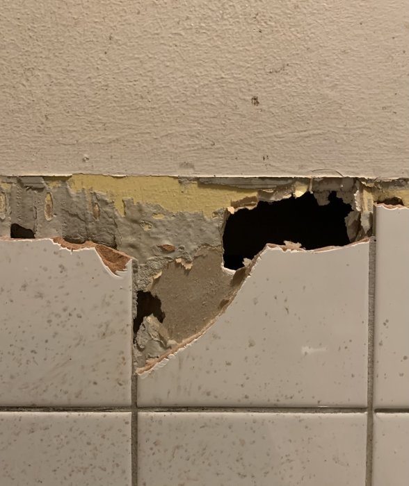 Skadad gipsvägg med delvis borttaget kakel som visar underliggande skador och träreglar.