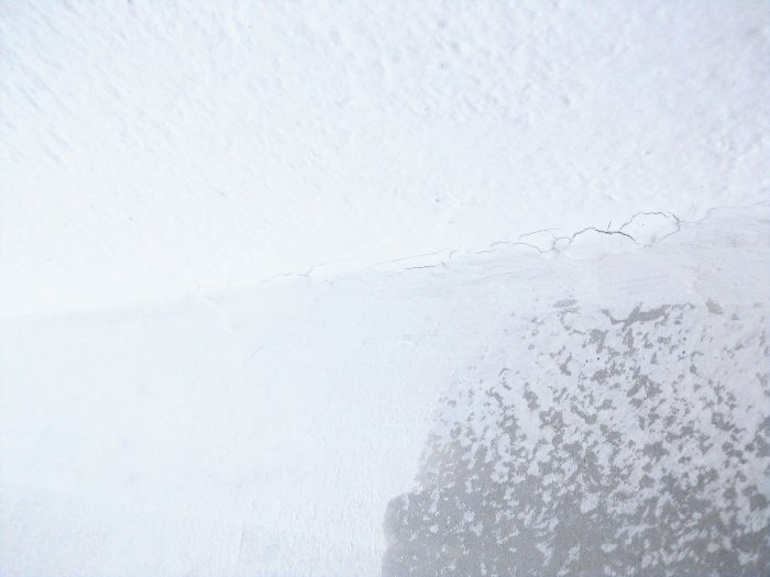 Sprickor i vit färg längs med fog där en vägg möter taket, antydan till underliggande tapetgrund.