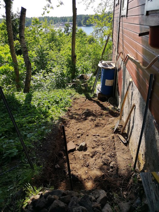 Utgrävd och jämnad mark vid stuga för tillbyggnad av kök, med skog och sjö i bakgrunden.