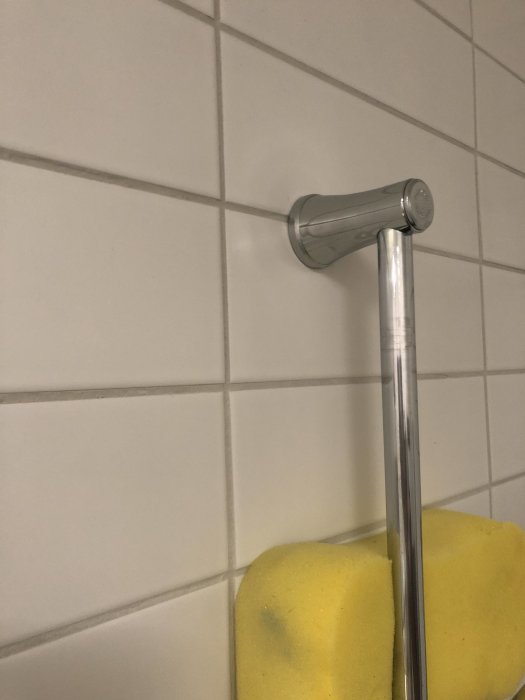 Duschstångshållare monterad på vit kakelvägg ovanför en gul svamp.