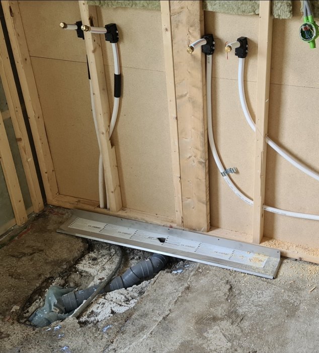 Inredning av ett byggprojekt med exponerade vattenrör och mätare på en trävägg, över ett upphackat golv för golvvärmeinstallation.