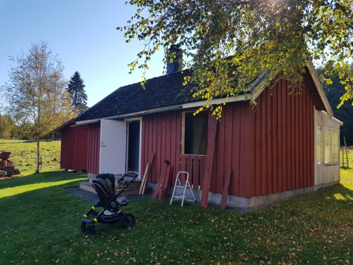 Rödmålad stuga med ny panel över gammalt fönster och underbrädor vid nytt fönster, barnvagn framför huset.