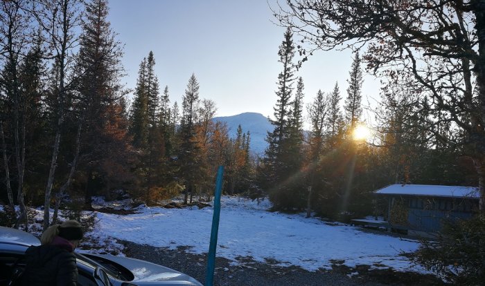 Person blickar mot en snötäckt tomt med träd och fjäll i bakgrunden under en solnedgång.