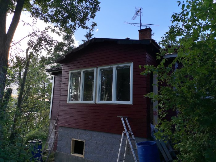 Exteriör bild före renovering med nya fönster på en röd träfasad, stege till vänster, omringad av grönska.