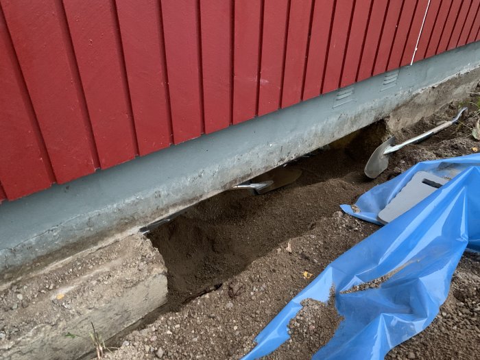 Hålrum i husgrund med synligt berg och sand under rödmålad panelvägg, bredvid grävverktyg och blå presenning.