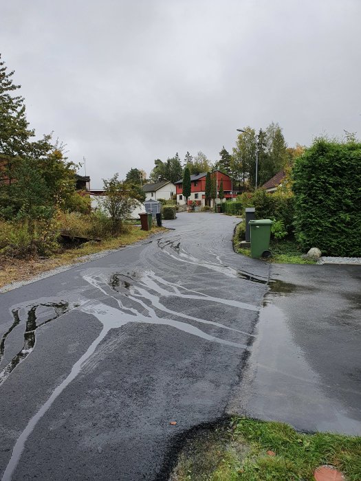 Ny asfalterad väg lutande mot garageuppfart med vattenansamling och hus i bakgrunden.