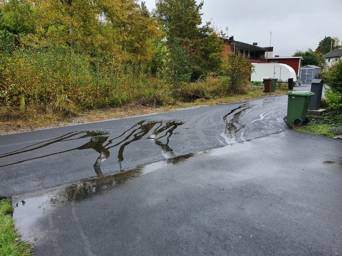 Ny asfalterad väg som lutar nedåt mot en garageuppfart, vattenspår visar avrinning mot uppfarten.