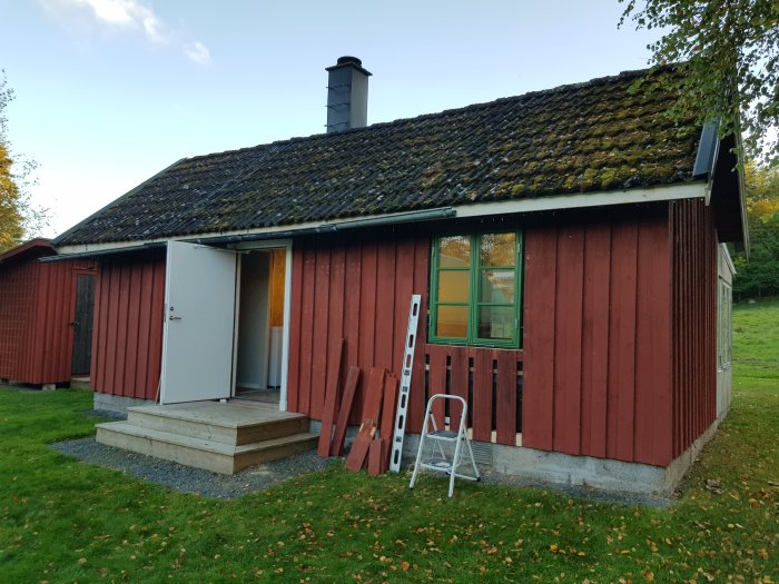 Rödmålad stuga med nyinstallerat fönster, stege lutad mot väggen, omgiven av höstlöv.
