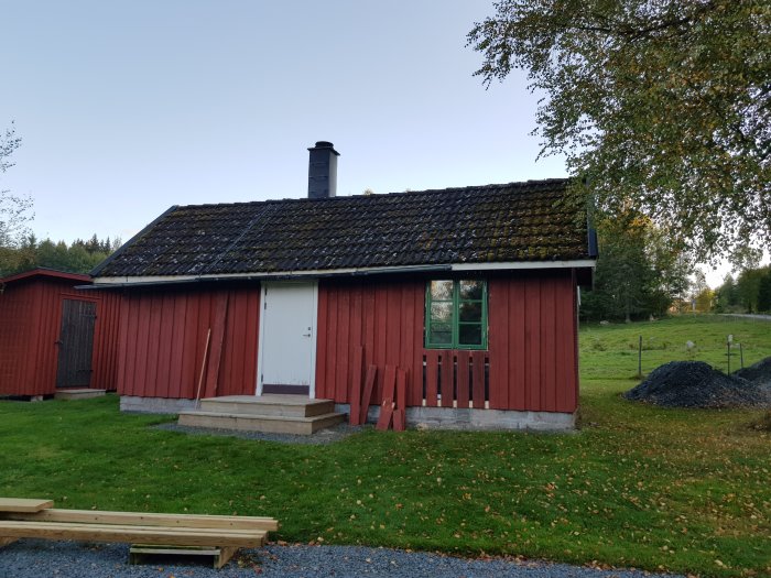 Rött trähus med nymonterat fönster, mossigt tak och byggmaterial på marken.