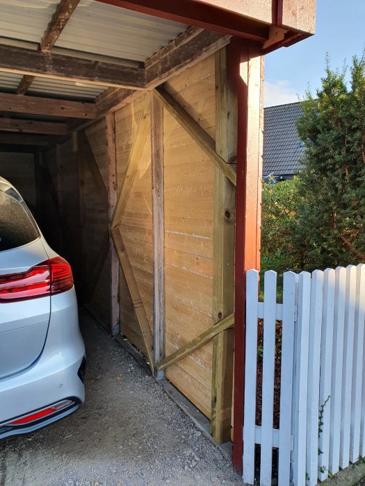 Nybytt träpanel på insidan av en carport med synlig stomme, intill en parkerad bil och ett vitt staket.