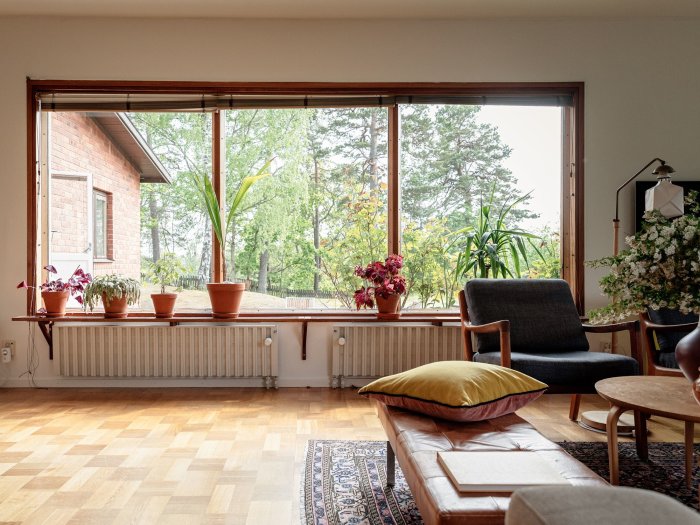 Interiör av ett rum med stora fönster som ser ut på en skog, med blomkrukor på fönsterbrädan och en mysig sittgrupp.
