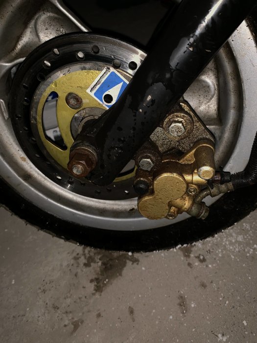 Närbild på en mopedens skivbroms och bromsok med synliga slitagespår och smuts.