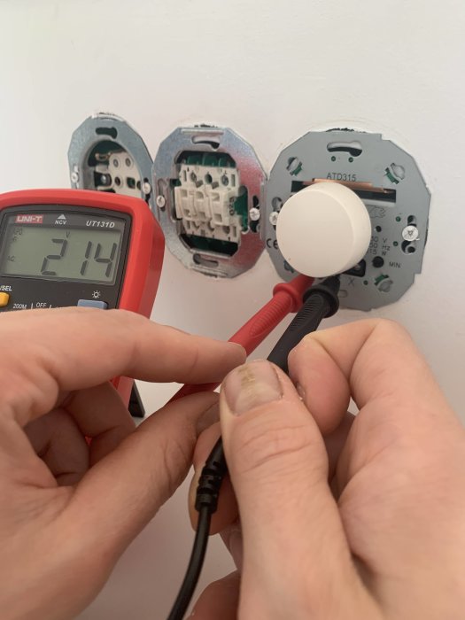 Multimeter visar 214 V på en strömbrytare, med osynliga spotlights och en dimmer i bakgrunden.