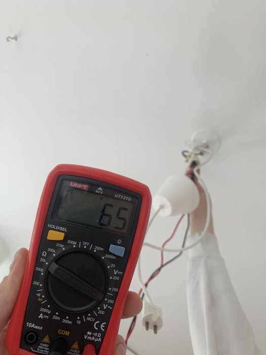 Hand håller en multimeter som visar 65 Volt vid ett lamputtag, med okopplade kablar och ej installerad lampa i bakgrunden.