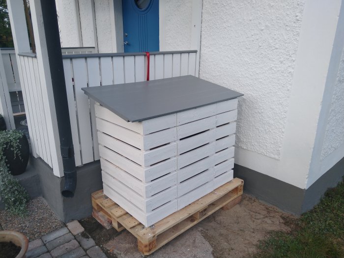 Hemmagjord vedförvaring i vitt placerad vid husvägg bredvid en veranda.