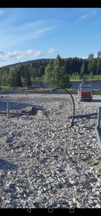 Byggarbetsplats med grus och synliga rörledningar i marken, grävmaskin i bakgrunden, omgiven av natur.