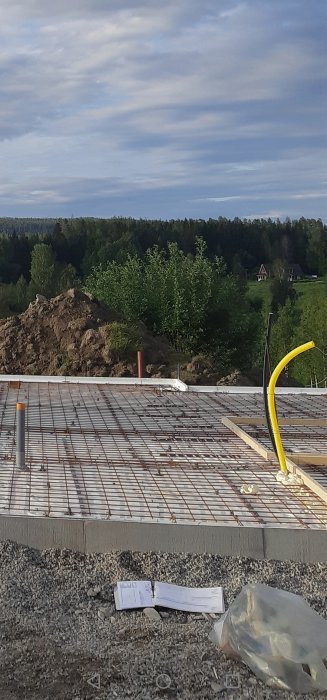 Konstruktionsbild av armerat betongfundament med tegelplan och grus i förgrunden, skogen i bakgrunden.