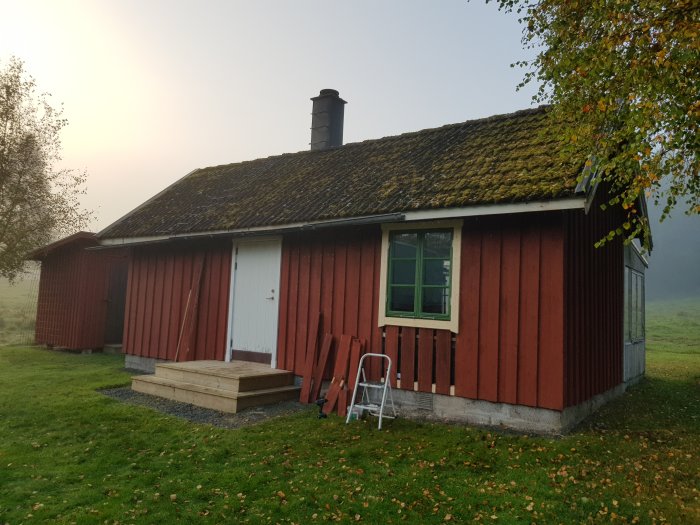 Rödmålad stuga med grundmålade fönsterfoder, lockbrädor och färdigmonterat fönster under en disig himmel.