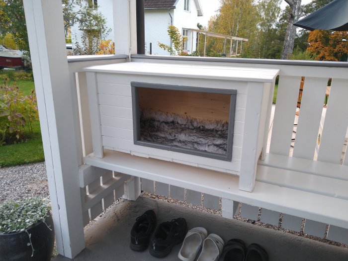 Vitmålad liten stuga med mjuk inredning för katt, placerad under en veranda.