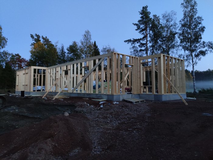 Färdigställda träytterväggar på ett husbygge i skymningen, med skog i bakgrunden och byggmaterial på marken.