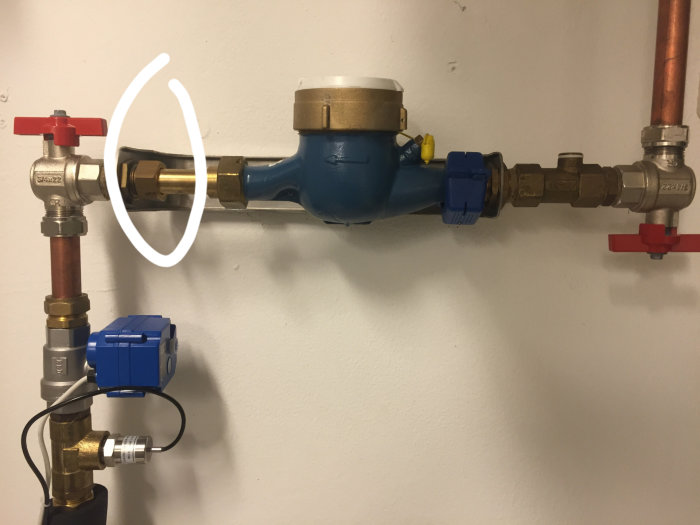VVS-installation med blå vattenmätare och röda avstängningsventiler på vägg, cirkeln markerar en stor mutter.