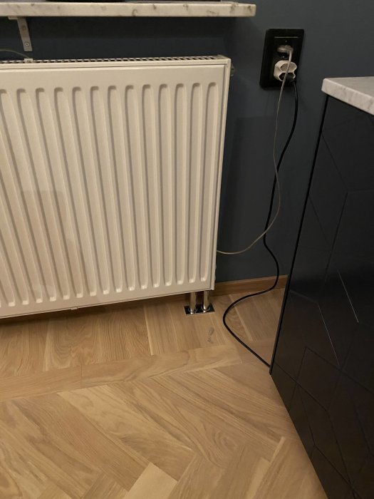 En vit radiator ansluten till kopparrör på ett trägolv nära en svart strömkontakt och en vägg med mörkgrå färg.