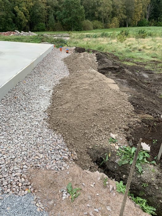 Lastad hög med 0/35 grus vid husgrund, grävmaskin arbete synligt, grön omgivning i bakgrunden.